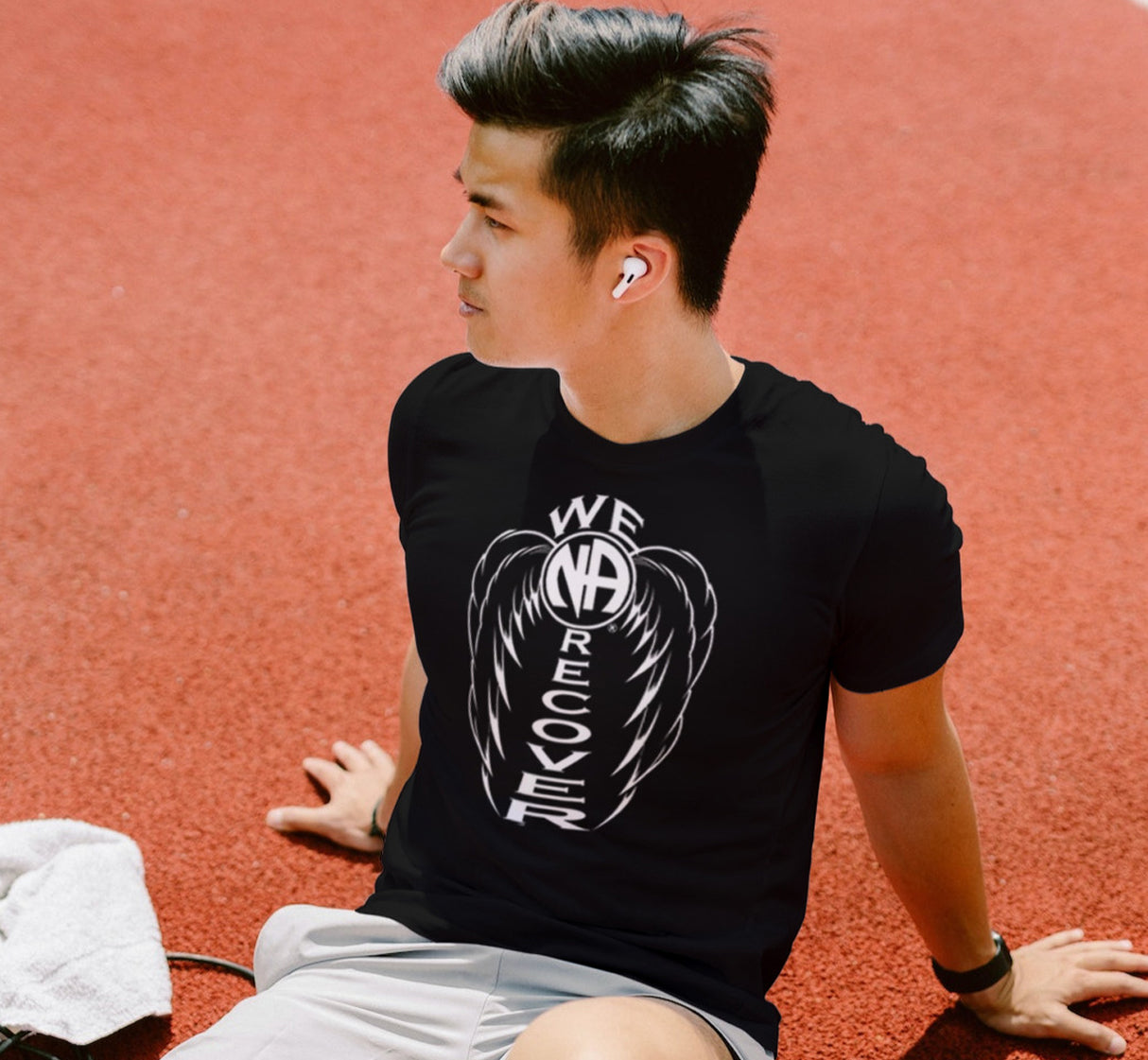 A person sitting on a track wearing a black T-shirt with 'WE RECOVER' text and wings design in white.