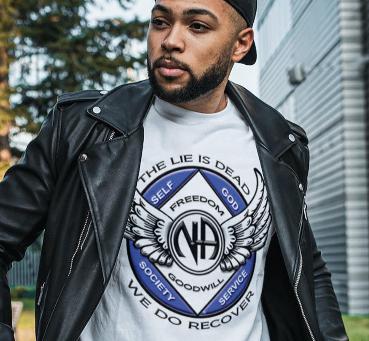A man wearing a white crew neck T-shirt with a graphic design, featuring a pair of wings and the Narcotics Anonymous emblem, paired with a black leather jacket and a black cap.
