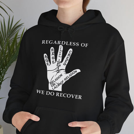 Person wearing a black hoodie with a white graphic and text, standing indoors next to a plant.