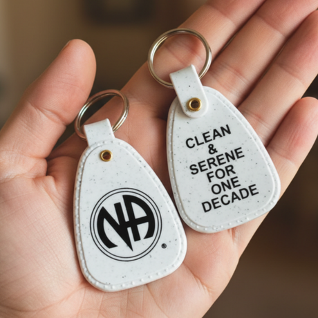 A hand holding two white guitar-pick-shaped keychains. One keychain has a black circular 'NA' logo, and the other has the text 'CLEAN & SERENE FOR ONE DECADE' in black.