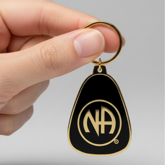 A hand holding a black teardrop-shaped keychain with a gold-tone metal ring and a gold circular logo featuring the letters 'NA'.