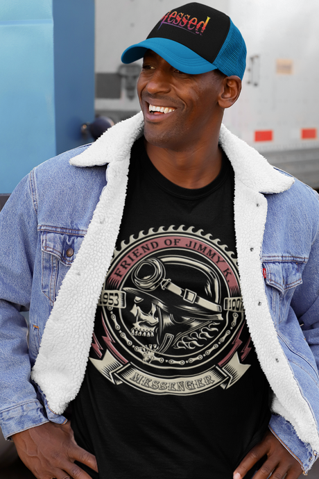 Man wearing a black t-shirt with a graphic design, blue denim jacket, and cap standing in front of a truck.