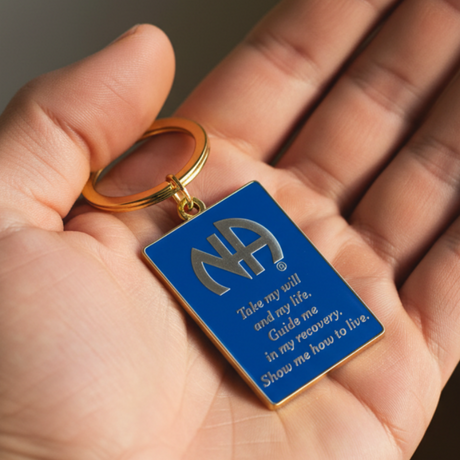 A close-up of a hand holding a blue rectangular keychain with a gold-tone metal ring and gold-tone lettering. The keychain features a large 'NA' emblem and the quote, 'Take my life and joy of it.