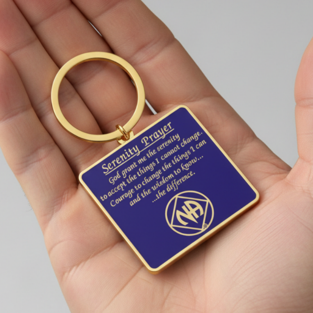 A square keychain with a gold-tone metal ring and border, featuring a deep blue face with white text reading 'Serenity Prayer' and a white circular symbol. The keychain is held in a person's hand against a neutral gray background.